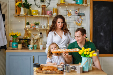 Happy family. A little blonde girl and her father with a French baguette are sitting at a table in the kitchen at home. At the same time, they bite the bun.