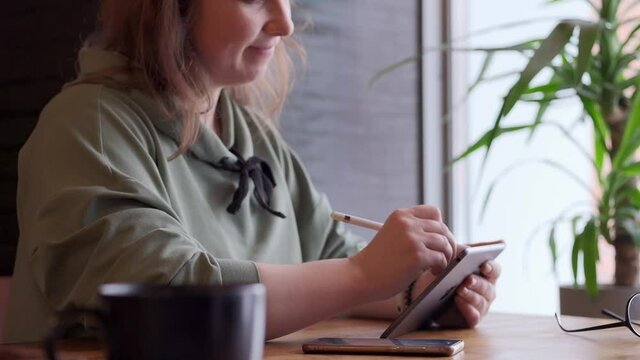 Long-haired woman draws on a tablet at cafe table in front of large window. Slow motion, side view