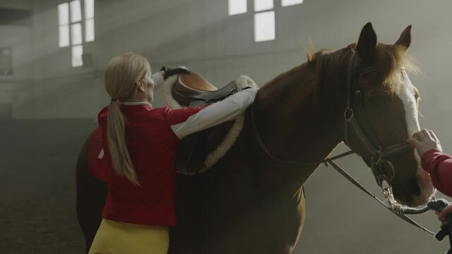 Jockey Put on and Removing Saddle from Horse Back. Ranch Worker Woman Preparing Thoroughbred Stallion for Race and Stroking. Girl with Horseback Equipment Tracking Shot Slow Motion