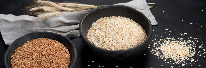 Rolled oat flakes in black bowls and golden wheat ears on dark background.