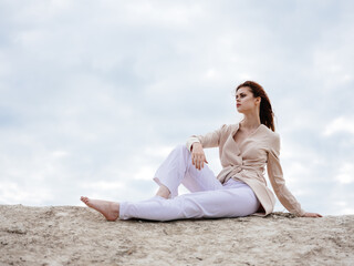 A woman in light clothes sits on the sand near the ocean in nature