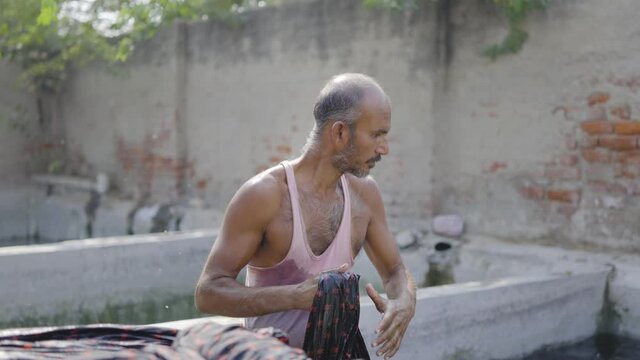 Shot of a middle-aged Indian male daily wage laborer or washerman of local or domestic textile dyeing firm is washing the dyed or colored long cotton sheet in the water 