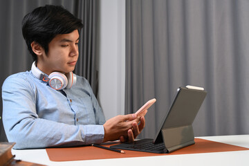 Casual asian man with headphone sitting in front of his computer tablet and using smart phone.