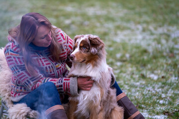 Tricolor Australian Shepherd sits next to a young woman by the campfire. In winter, snow on the grass. The fire is burning at dusk. Hugging the dog. Selective focus