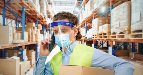 Man in protective medical mask standing in warehouse and holding cardboard box and talking on mobile phone.