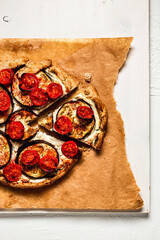 A close-up of a slice of a galette or tart with ricotta cheese, aubergine and cherry tomatos on baking paper and white wooden board, view from above