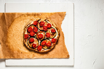 A galette or tart with ricotta cheese, aubergine and cherry tomatos on baking paper and white wooden board, view from above, copy space