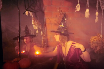 Blonde witch in red dress and black hat with candle and mirror in Halloween decoration indoors.