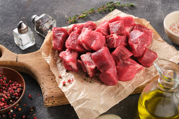 Raw ingredients for goulash. Fresh raw chopped beef on an wooden board, spices, herbs and vegetables on dark grey background