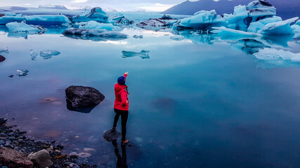 A girl in pink jacket standing on the rock in a shallow water. She is pointing at the drifting ice bergs in glacier lagoon. Glacier has many shades of blue. Global warming causing melting of glaciers  © Chris