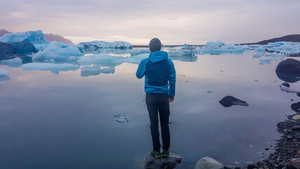 Young man in a jacket standing on the rock in a shallow water. He looks at the lagoon, with one hand on his face. Glacier lagoon full of drifting ice bergs. Global warming causing melting of glaciers  © Chris