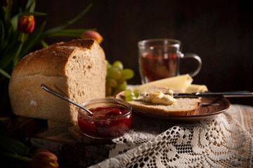 Frühstück ist fertig! Brot ist geschnitten, mit Butter geschmiert, Glas Tee , Weintrauben, Tulpen und Erdbeere Marmelade  stehen daneben auf dem Holztisch  Mit weißen Tischdecke dekoriert. Rustic