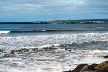 Fototapeta premium Lahinch town coast line. County Clare, Ireland, Sunny day, blue cloudy sky. Nobody. Powerful waves moving towards the beach. Surfer in dark wet suit in the water