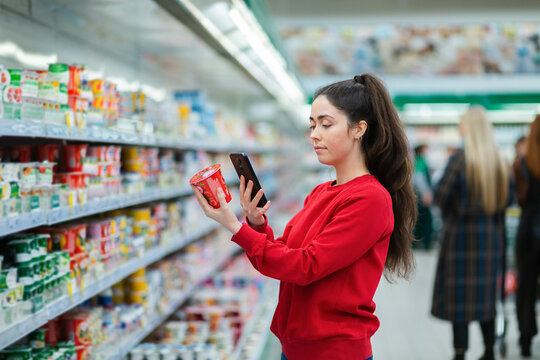 A young woman scans the QR code on a package of yogurt. In the background, a supermarket with visitors in a blur. The concept of modern technologies