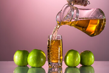 Fresh apple juice is poured from a carafe into a glass. Nearby are green apples on the reflective surface of the table. Pink background. Summer mood