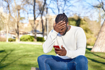 Fototapeta premium African American man sitting on a park bench looking at his cell phone. Concerned young man. Concept of technology