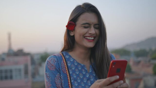 A young Indian Asian cheerful attractive woman is holding a mobile phone in hands and watching a comedy video or movie on an online stream or OTT platform smiling and standing outdoors in the evening