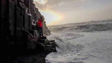 A girl wearing pink jacket standing at the basalt columns, when the tide washed in at Reynisfjara Black Sand Beach, Iceland. Power of the sea, unexpected wave. Sun setting behind the basalt wall.