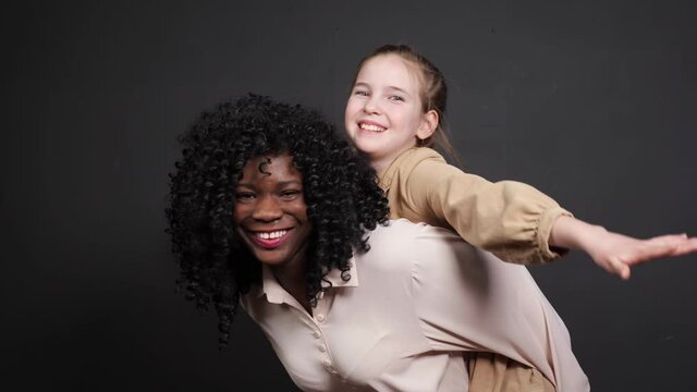 Stylish black stepmother in white blouse holds on backs adopted schoolgirl with fair hair raising hands and smiles cheerfully posing at home
