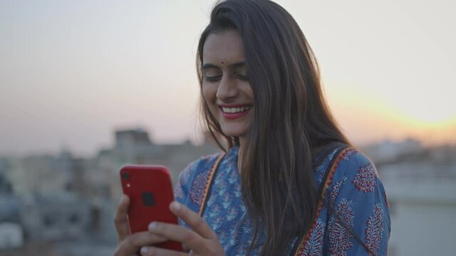 A young Indian Asian cheerful attractive woman is holding a mobile phone in hands and watching a comedy video or movie on an online stream or OTT platform smiling and standing outdoors in the evening