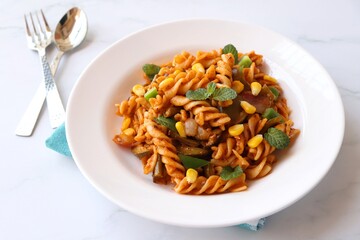 Whole wheat fusilli pasta with vegetables. Vegetable Pasta with zucchini, mushrooms, corn, and peppers on a white plate. Garnished with mint leaves. on a white marble table with cutlery. copy space.