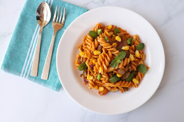 Whole wheat fusilli pasta with vegetables. Vegetable Pasta with zucchini, mushrooms, corn, and peppers on a white plate. Garnished with mint leaves. on a white marble table with cutlery. copy space.