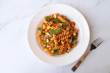 Whole wheat fusilli pasta with vegetables. Vegetable Pasta with zucchini, mushrooms, corn, and peppers on a white plate. Garnished with mint leaves. on a white marble table with cutlery. copy space.
