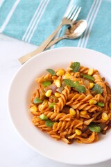 Whole wheat fusilli pasta with vegetables. Vegetable Pasta with zucchini, mushrooms, corn, and peppers on a white plate. Garnished with mint leaves. on a white marble table with cutlery. copy space.