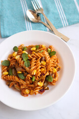 Whole wheat fusilli pasta with vegetables. Vegetable Pasta with zucchini, mushrooms, corn, and peppers on a white plate. Garnished with mint leaves. on a white marble table with cutlery. copy space.