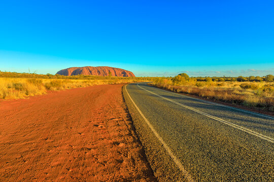 Road Leading To Uluru Ayers Rock At Vibrant Color Of Sunset. The Huge Sandstone Monolith Icon Of Australian Outback In Uluru-Kata Tjuta National Park. Central Australia, Northern Territory. Copy Space