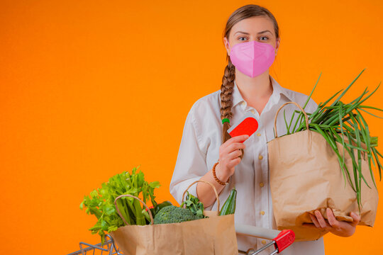 Attractive young lady wearing an pink CORONAVIRUS MASK (COVID19) close to a shopping cart having shopping bags in it with fresh veggies showing her fidelity card.