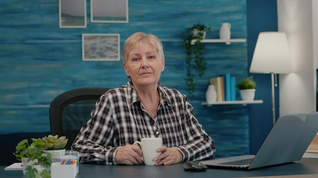 Senior focused woman looking at camera sitting in workplace at desk working from home while old husband watching tv in background. Retired business person prepared for online meeting using moden
