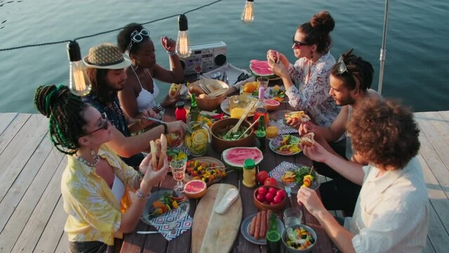 High angle tilt down shot of company of young multiethnic male and female friends sitting at dinner table, eating food and speaking at lake party on pier on sunny day