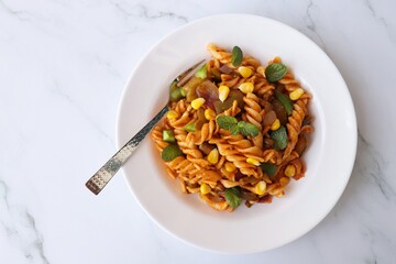 Whole wheat fusilli Pasta Arrabbiata with vegetables. Vegetable Pasta with zucchini, mushrooms, corn, and peppers on a white plate. on a white marble table with cutlery. copy space.