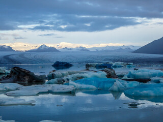Obraz premium Beautiful glacier lagoon. Thousands of icebergs drifting lazily towards the sea, shining in many shades of blue. Soft sunset in the back. Thick clouds above the lagoon. Glacier's cap in the back