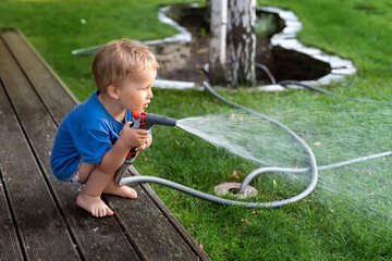 Cute adorable caucasian blond toddler boy enjoy having fun watering garden flower and lawn with hosepipe sprinkler at home backyard at sunny day. Child little helper learn gardening at summer outdoor
