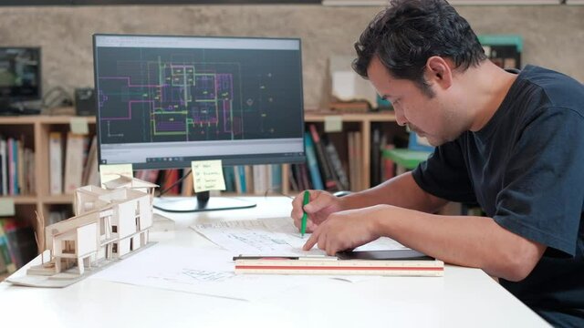 An Asian architect in a dark shirt is working on a design in his studio using a computer-aided design program. And scribble down on the construction plan.