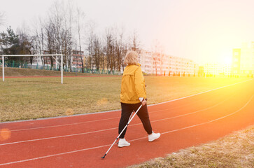 An elderly woman in a yellow sports jacket practices Nordic walking outdoors on the stadium's rubber treadmill. A sunny sunset. Pensioners healthy lifestyle