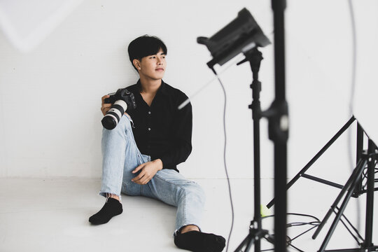 Asia young handsome successful male photographer wears black long sleeve shirt hold DSLR camera in hand while sit on floor leaning on white background checking photo from album in indoor photo studio