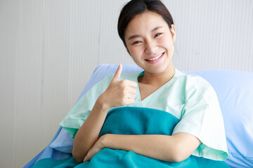 Asian young long hair happy female patient wears green hospital uniform covered by blanket sit lean on blue pillow on bed hold hand shows thumb up when recovery and in good condition in private room