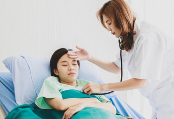 Obraz na płótnie Canvas Asian long brown hair female doctor visit patient in green hospital uniform covered by blanket lay down on blue pillow bed check temperature by touching forehead and listen beat pulse by stethoscope