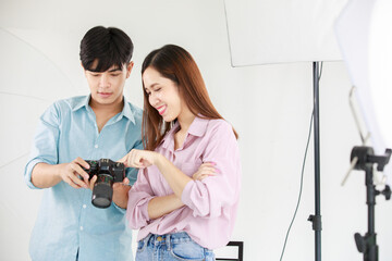 Asian young beautiful long brown hair female model and handsome male photographer holding full frame dslr camera in hands stand smiling talking together checking shooting image from screen in studio
