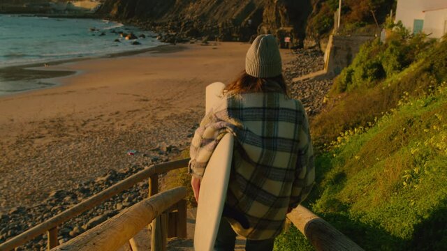 Handheld natural shot of authentic casual young female surfer. Woman with surfboard walk down stairs to beautiful epic surfing beach at sunset. Follow freedom and wanderlust lifestyle. Cinematic vibe