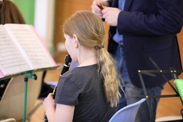 A little girl with long blonde hair pulled back in a ponytail in the classroom in music class looks at the notes and plays the clarinet to the teacher standing next to her. School education concept
