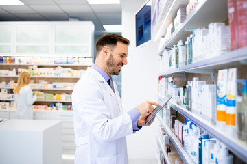 Pharmacist standing by the shelves with medicines and typing on tablet in pharmacy store.