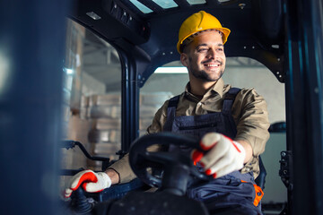 Young caucasian male in working uniform and yellow hardhat operating forklift machine in warehouse storage room. - obrazy, fototapety, plakaty