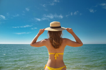 a slender young woman in a swimsuit and a hat stands waist-deep in the sea water on a sunny day. Holds his hat with his hands. Back view
