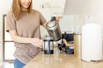 Happy cropped young woman making coffee in kitchen - french press coffee. Caucasian female model in...