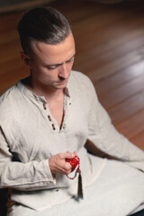 A young man in gray practice clothes sits in a lotus position with a red rosary in his hand in meditation. Dark room calmness of mind and body.