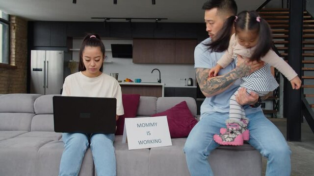 Busy serious pretty asian businesswoman networking online on business project using laptop, ignoring handsome husband babysitting with cute toddler daughter and recommending to call 911 at home.